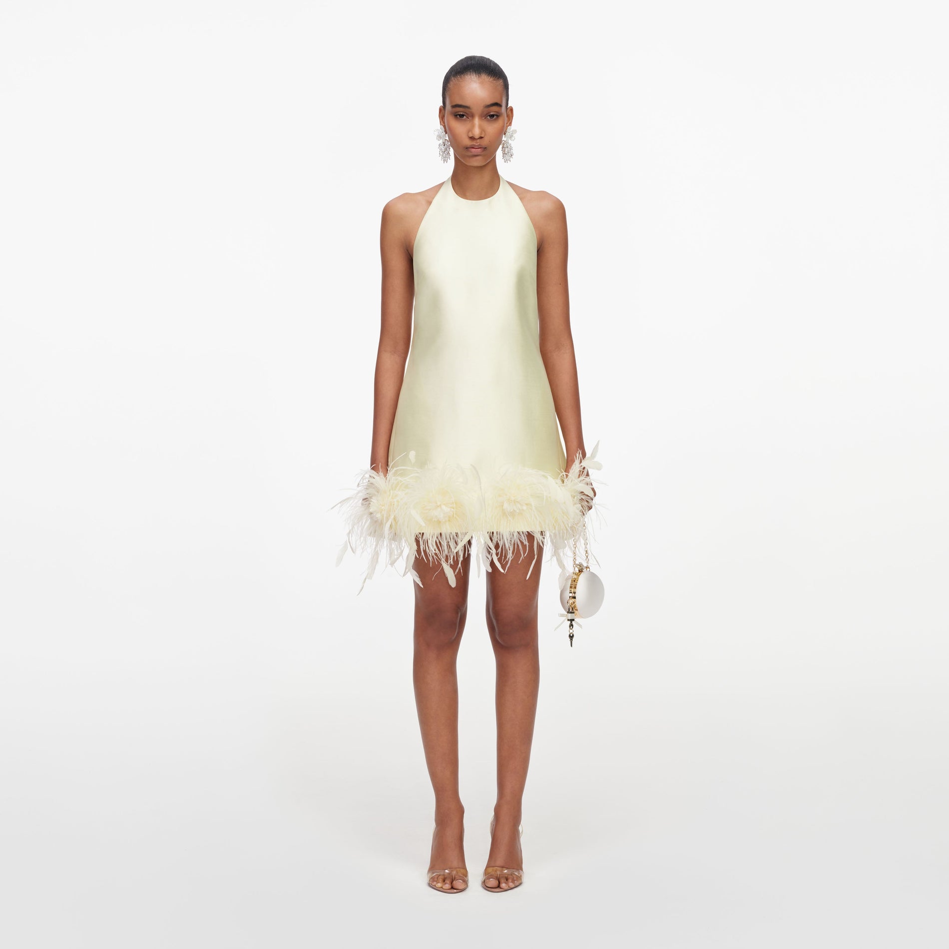 Model wearing a pale yellow halter neck mini dress with feather trim and a white spherical handbag