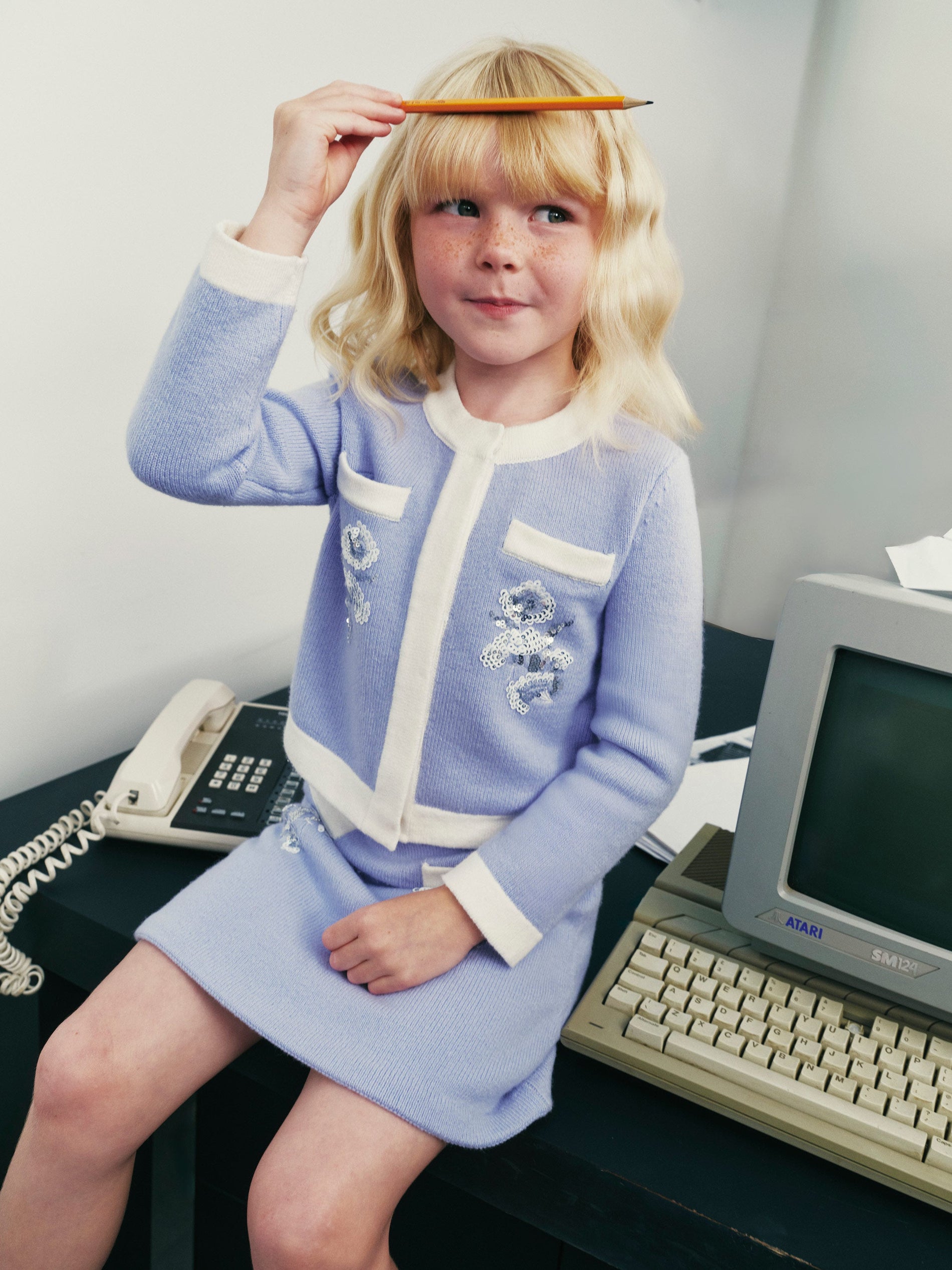 Child in a light blue cardigan and skirt set, holding a pencil on their head, next to a vintage computer