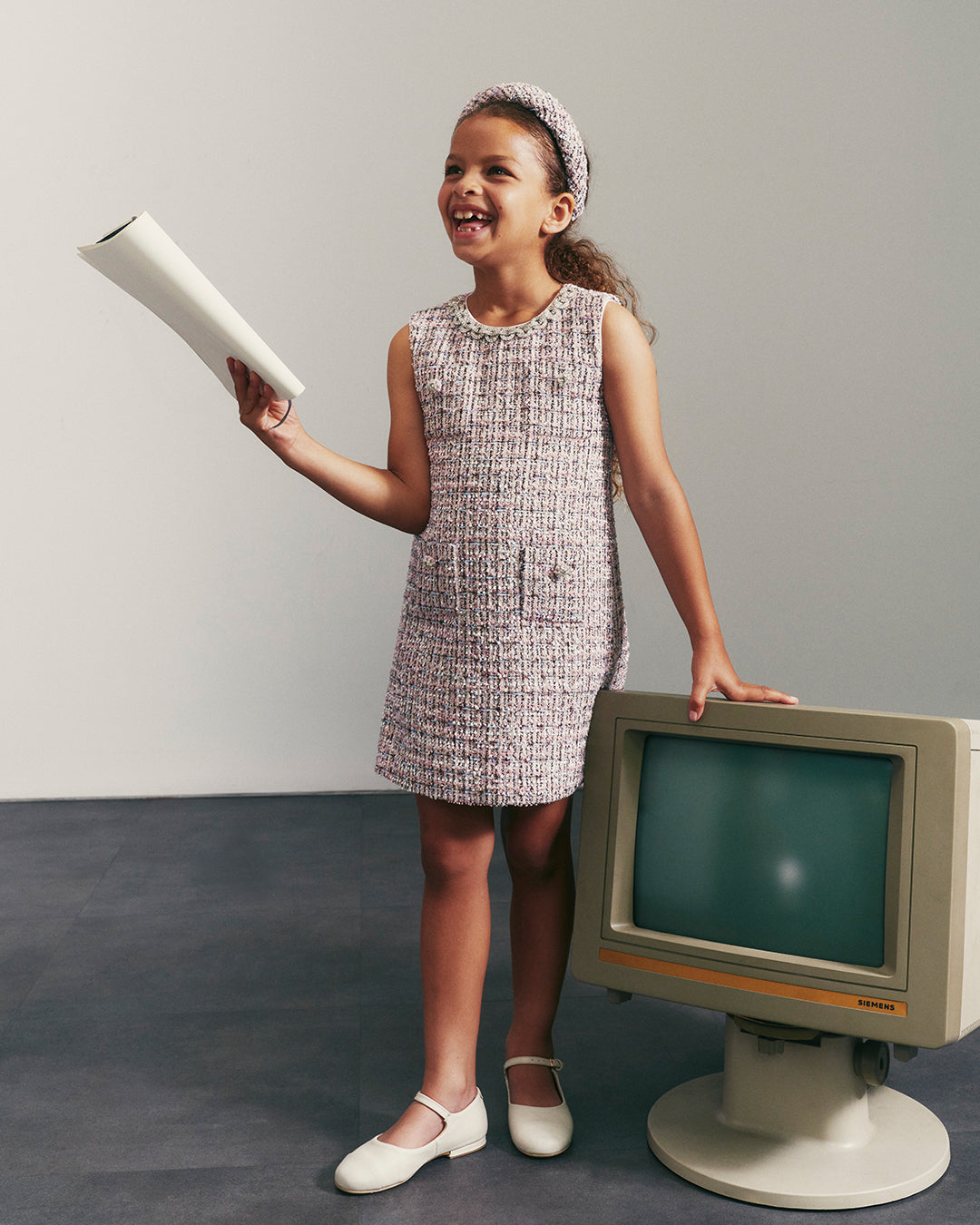 Girl in tweed dress and headband holding paper, standing next to a vintage computer monitor