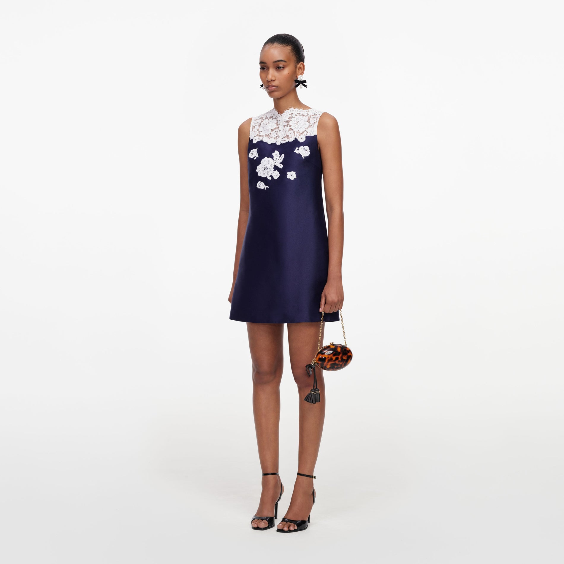 Woman in navy blue dress with white lace trim and floral embroidery, black heels, and a tortoiseshell clutch