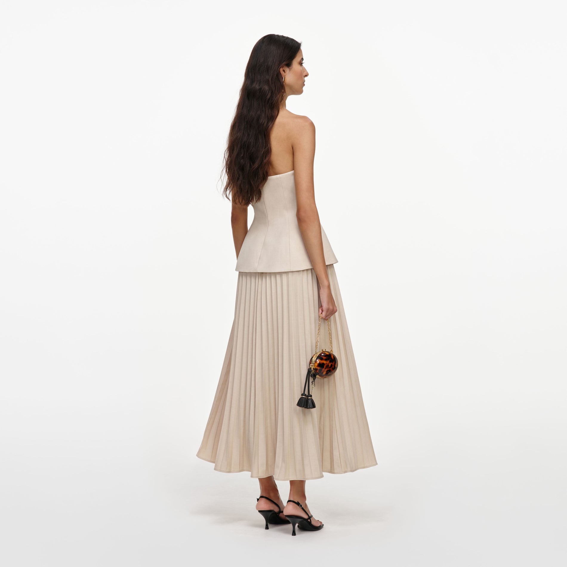 Woman in a strapless top and pleated skirt, holding a small round tortoiseshell bag with black tassels