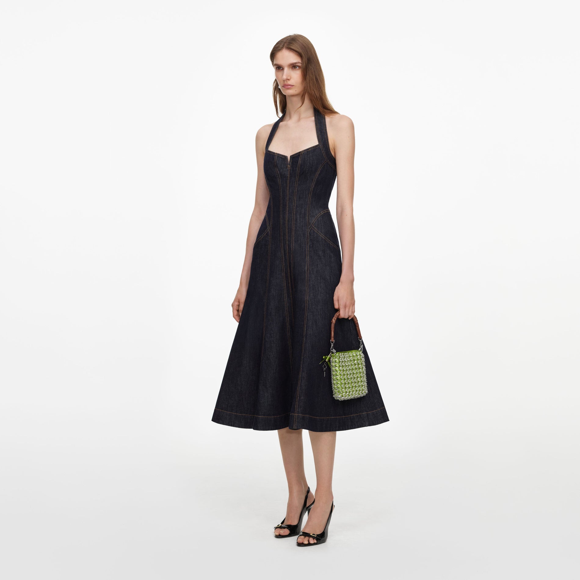 Model in a dark denim halter dress with a full skirt, holding a small green beaded handbag and wearing black slingback heels