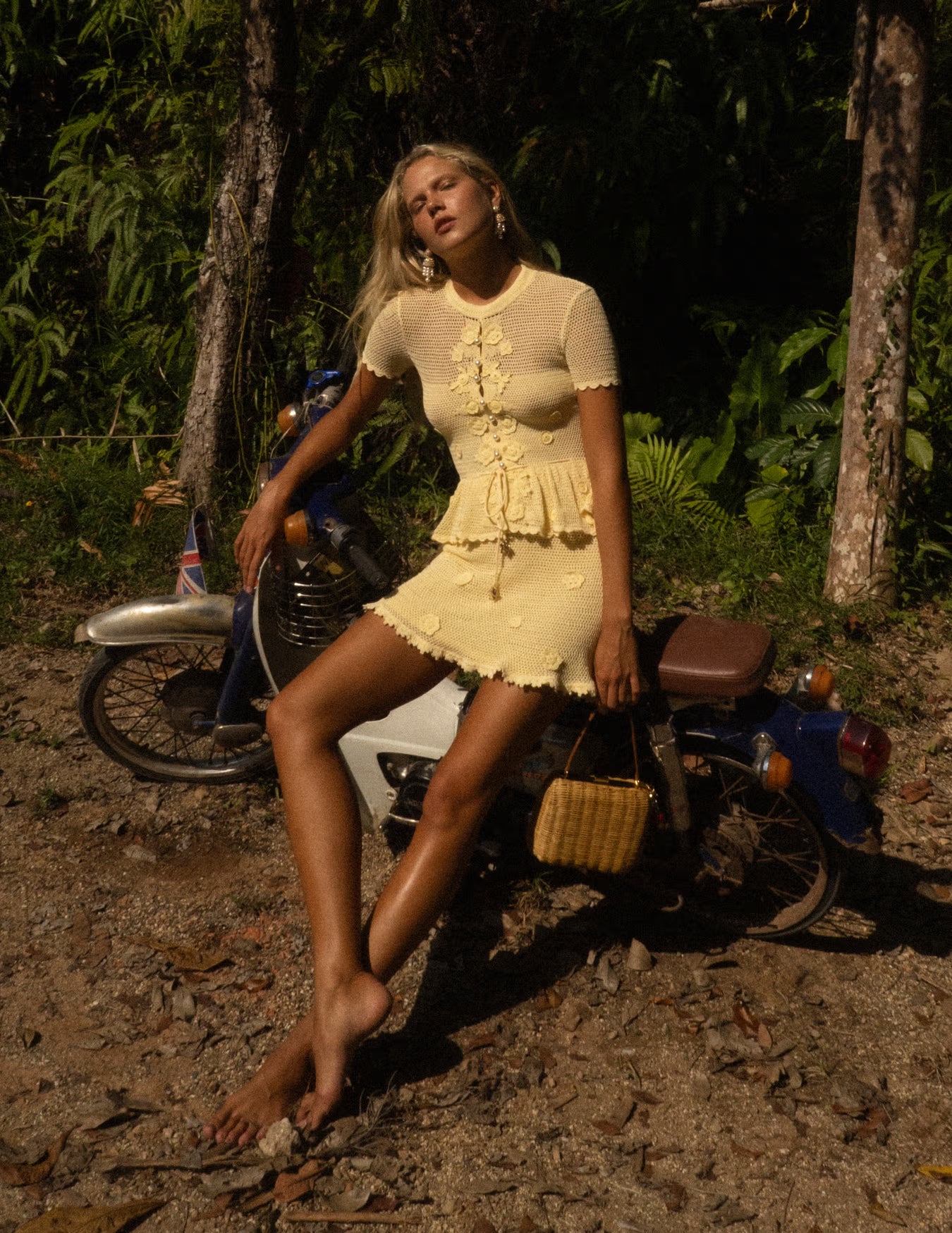 Woman in a yellow knit mini dress standing next to a motorcycle in a forest setting.